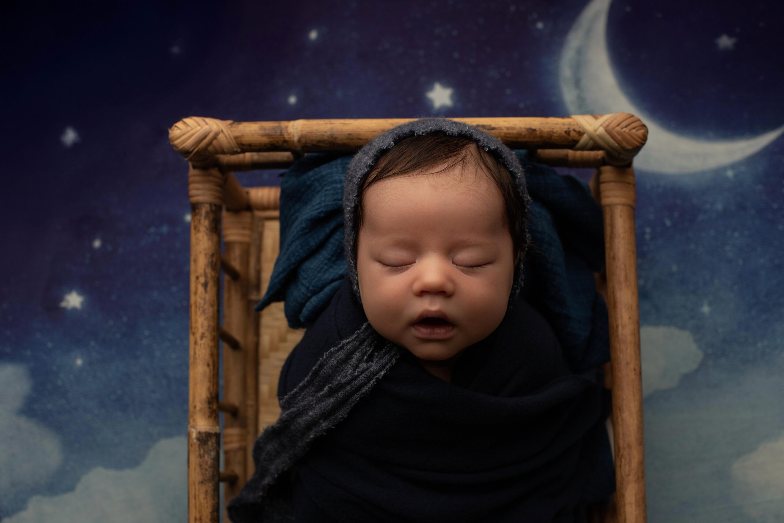Baby wrapped with blue fabric in a bed on a cloud backdrop.