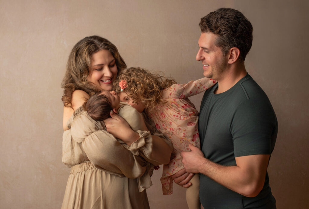 Family newborn session with older sister kissing baby and parents watching in awe.
